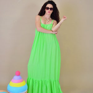 May include: A woman wearing a bright green maxi dress with a ruffled hem. She is wearing sunglasses and a gold necklace. The dress is sleeveless and has a loose fit.