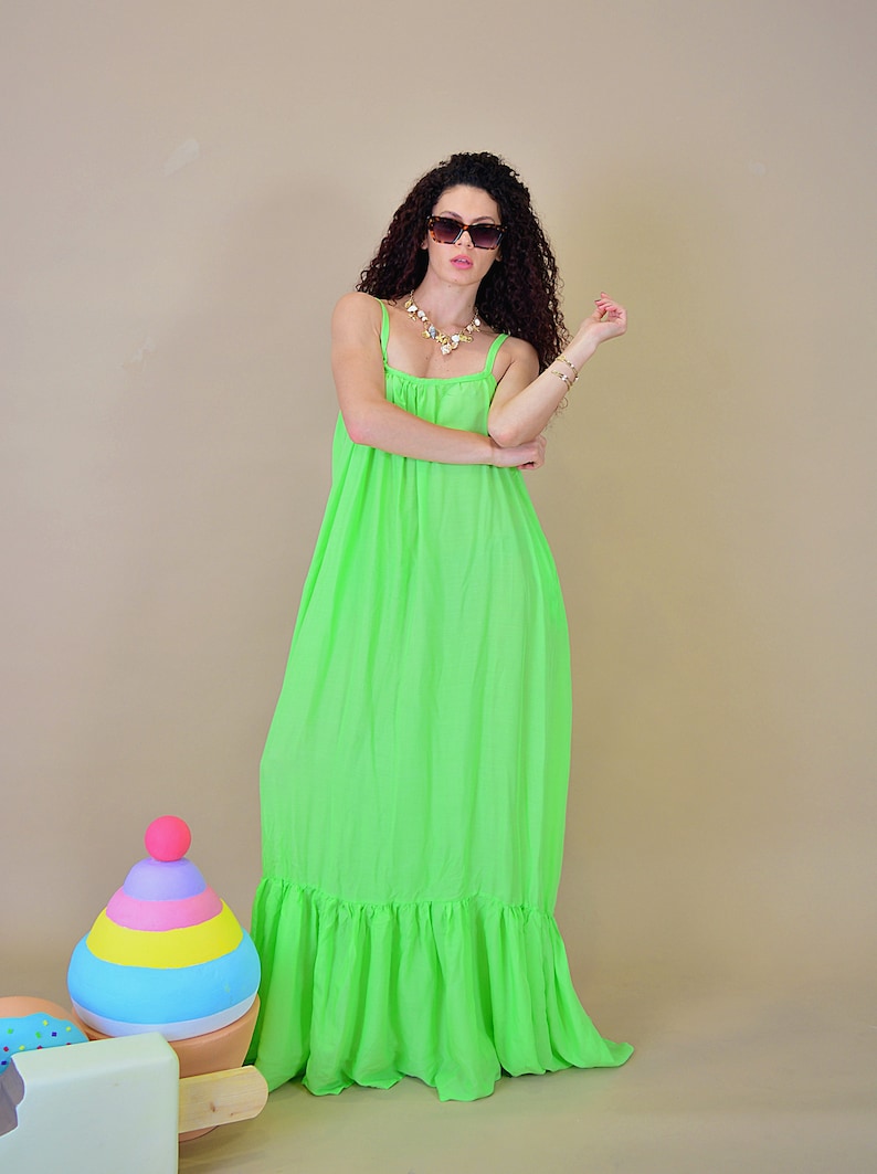 May include: A woman wearing a bright green maxi dress with a ruffled hem. She is wearing sunglasses and a gold necklace. The dress is sleeveless and has a loose fit.
