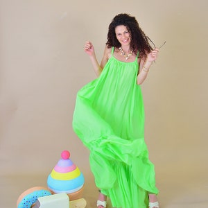 May include: A woman wearing a bright green maxi dress smiles and holds a pair of sunglasses. She stands in front of a colorful wooden toy that resembles a stack of ice cream cones and a popsicle.