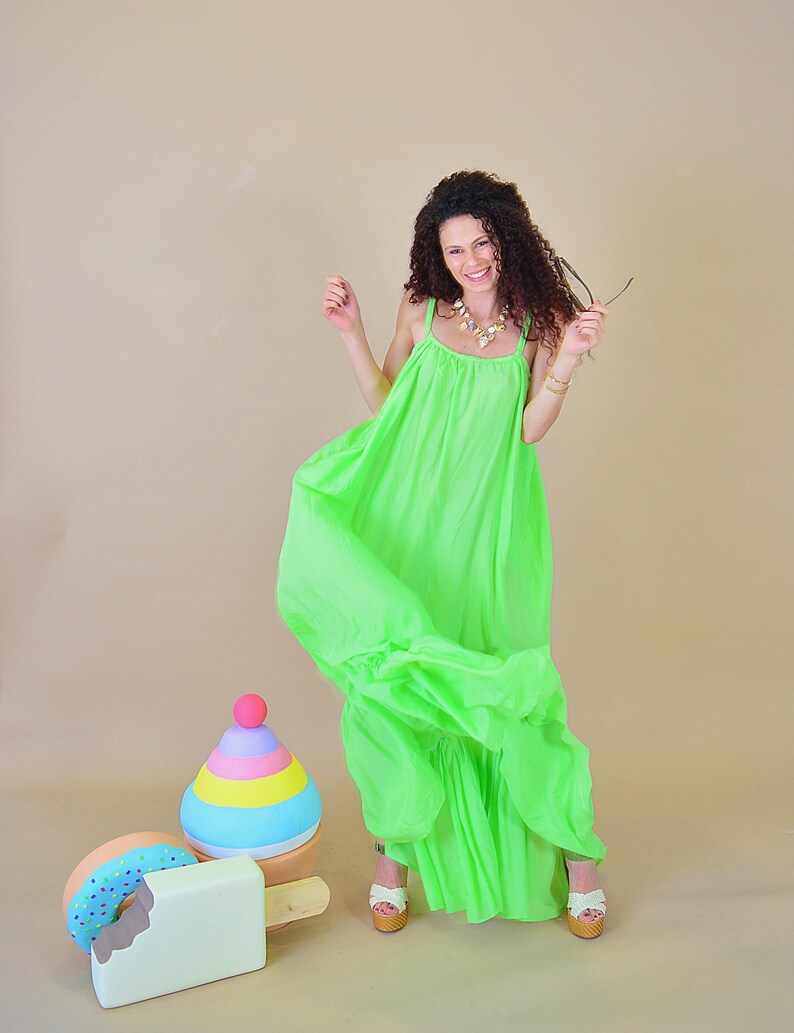 May include: A woman wearing a bright green maxi dress smiles and holds a pair of sunglasses. She stands in front of a colorful wooden toy that resembles a stack of ice cream cones and a popsicle.