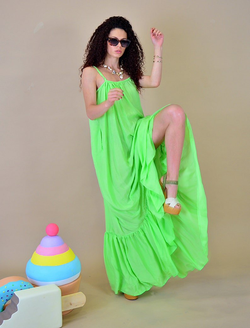 May include: A woman wearing a bright green maxi dress with a ruffled hem and white sandals. She is standing in front of a colorful toy ice cream cone and a donut.