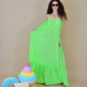 May include: A woman wearing a bright green maxi dress with a ruffled hem and sunglasses. She is standing in front of a light brown background with colorful toy props.