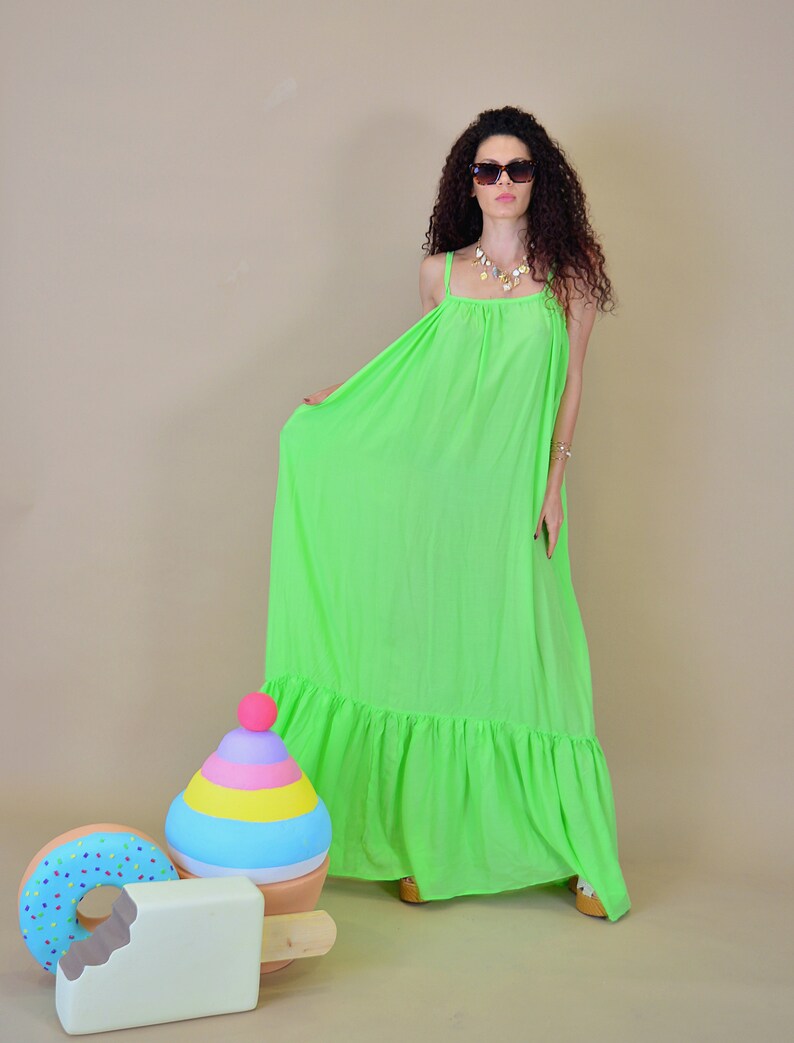 May include: A woman wearing a bright green maxi dress with a ruffled hem and sunglasses. She is standing in front of a light brown background with colorful toy props.