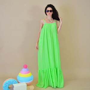 May include: A woman wearing a bright green maxi dress with a ruffled hem and sunglasses. She is standing in front of a colorful toy ice cream cone and a donut.