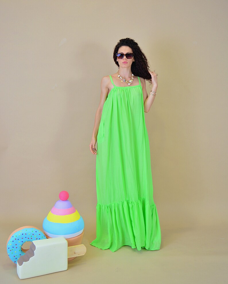 May include: A woman wearing a bright green maxi dress with a ruffled hem and sunglasses. She is standing in front of a colorful toy ice cream cone and a donut.