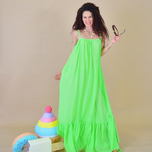 May include: A woman wearing a bright green maxi dress with a ruffled hem, standing in front of a light brown background. She is holding a pair of sunglasses in her right hand. There are three wooden toys in the foreground: a donut, a popsicle, and a colorful stacking toy.