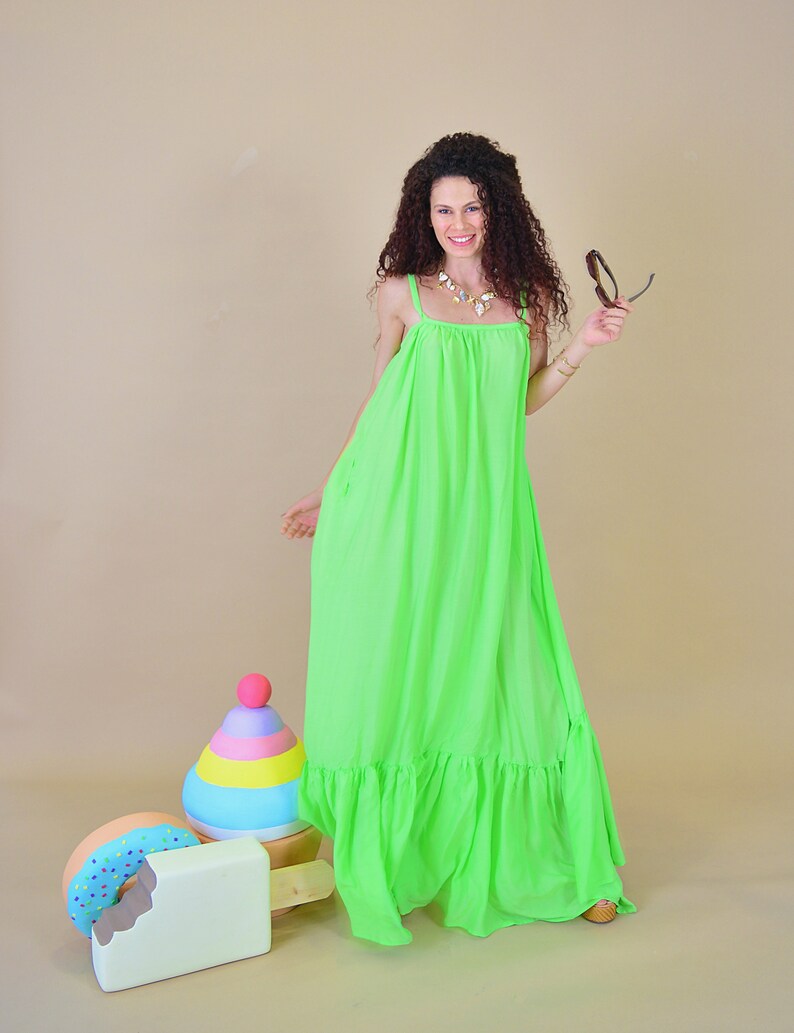 May include: A woman wearing a bright green maxi dress with a ruffled hem, standing in front of a light brown background. She is holding a pair of sunglasses in her right hand. There are three wooden toys in the foreground: a donut, a popsicle, and a colorful stacking toy.