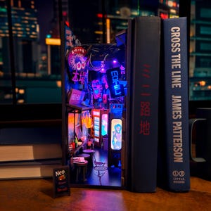Op de afbeelding: Een miniatuur diorama van een Japanse straatsc&egrave;ne, verlicht met neonlichten, geplaatst tussen twee boeken. De sc&egrave;ne bevat borden met Japanse tekst, een kleine tafel en een miniatuur menubord. De boektitels zijn "Cross the Line" en "路地".