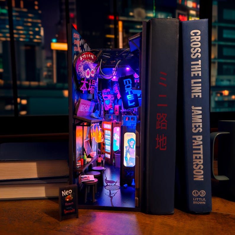 Op de afbeelding: Een miniatuur diorama van een Japanse straatsc&egrave;ne, verlicht met neonlichten, geplaatst tussen twee boeken. De sc&egrave;ne bevat borden met Japanse tekst, een kleine tafel en een miniatuur menubord. De boektitels zijn "Cross the Line" en "路地".