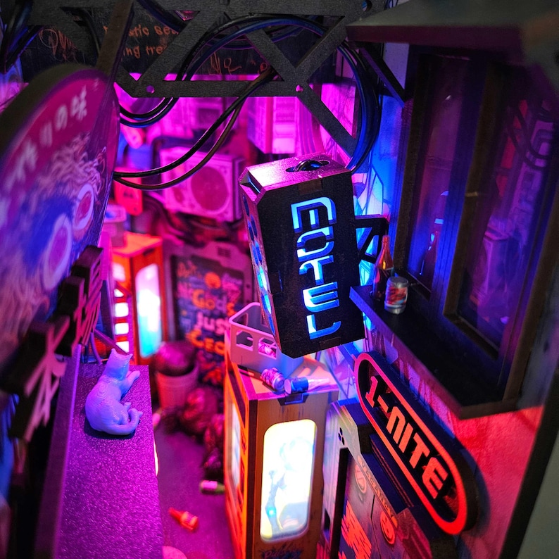 Op de afbeelding: Miniatuur diorama van een Japanse straat met neonverlichting. De sc&egrave;ne toont oplichtende borden, waaronder een met Japanse karakters en een ander met de tekst "1-NITE". Een blauw kattenbeeldje zit op een richel, verlicht door paars en blauw licht.