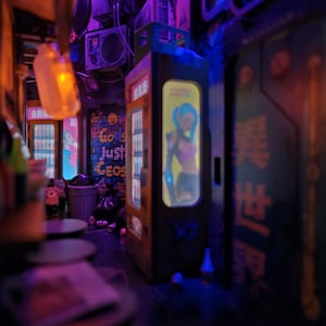 Op de afbeelding: Een miniatuur cyberpunk steeg met automaten, neonreclame en afval. De automaten tonen personages in anime-stijl. De sc&egrave;ne is verlicht met paarse en oranje tinten, wat een futuristische sfeer cre&euml;ert.