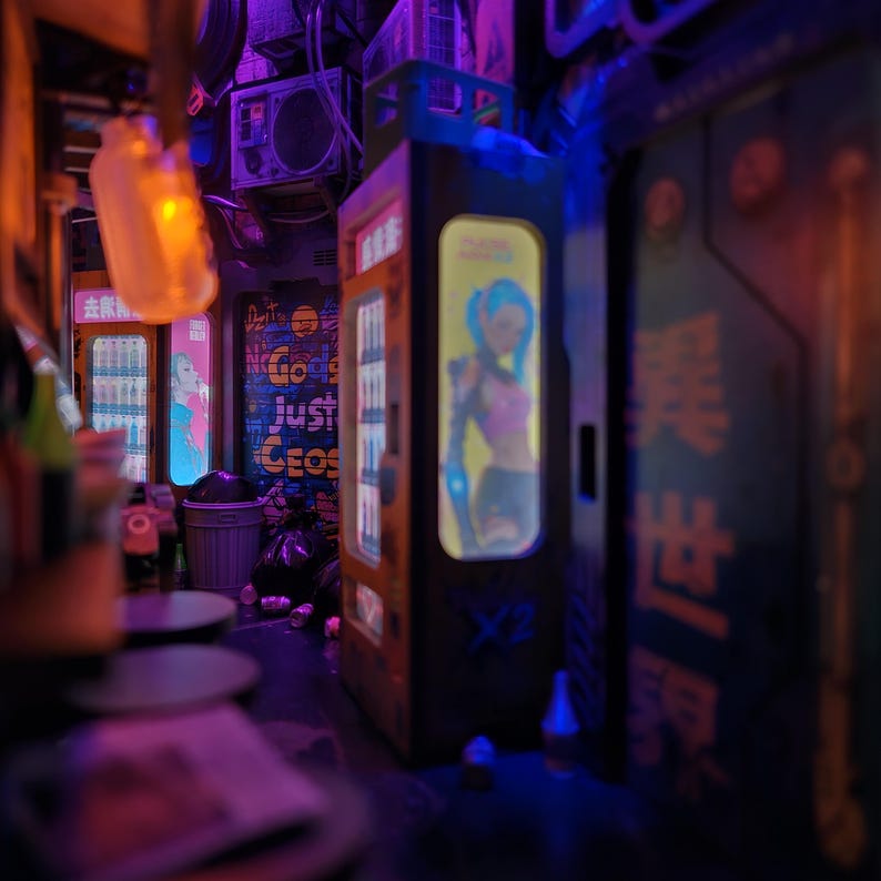 Op de afbeelding: Een miniatuur cyberpunk steeg met automaten, neonreclame en afval. De automaten tonen personages in anime-stijl. De sc&egrave;ne is verlicht met paarse en oranje tinten, wat een futuristische sfeer cre&euml;ert.