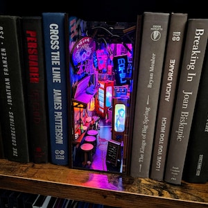 Op de afbeelding: Een unieke boekenplank-inzet met een miniatuur, verlichte Japanse straatsc&egrave;ne. Het diorama bevindt zich tussen hardcover boeken op een houten plank. De sc&egrave;ne bevat neonreclame, een bar en kleine tafels, wat een levendig beeld cre&euml;ert.