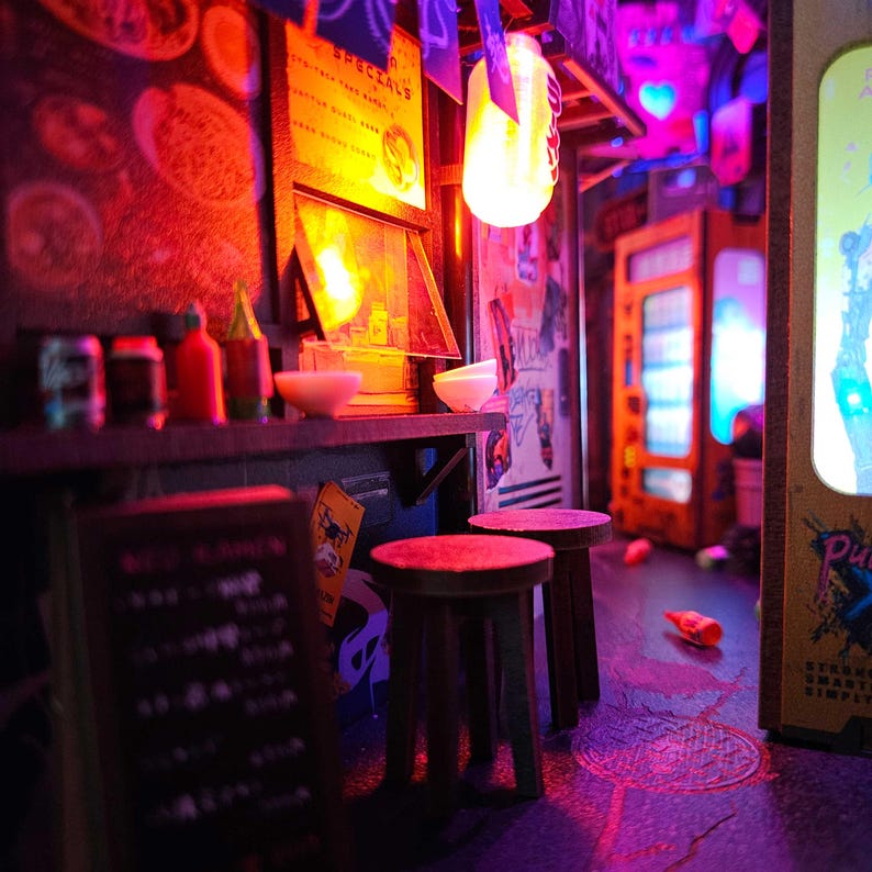 Op de afbeelding: Miniatuur diorama van een neonverlichte Aziatische straatvoedselkraam. De sc&egrave;ne omvat een bar met kommen en flessen, krukken en een automaat. De verlichting is een mix van rood, oranje en paars.