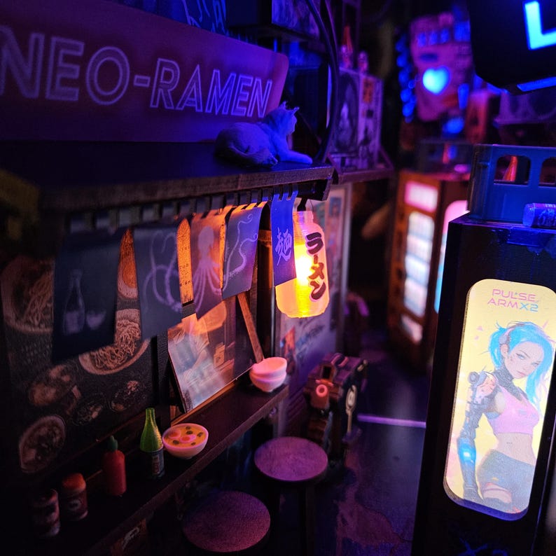 Op de afbeelding: Miniatuur cyberpunk ramenwinkel met neon "NEO-RAMEN" bord en een kattenbeeldje. Voorzien van hangende lantaarns, kleine tafels en posters. Een bord leest "PULSE ARMX2" met een illustratie van een vrouwelijk personage. De sc&egrave;ne wordt verlicht met blauwe en gele lichten.