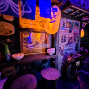 Op de afbeelding: Een miniatuur Japanse ramenwinkelsc&egrave;ne met een warme, uitnodigende gloed. De sc&egrave;ne omvat een kleine toonbank, krukken en posters. Er hangen lantaarns en een kleine robot. De winkel is versierd met Japanse karakters en voedselillustraties.