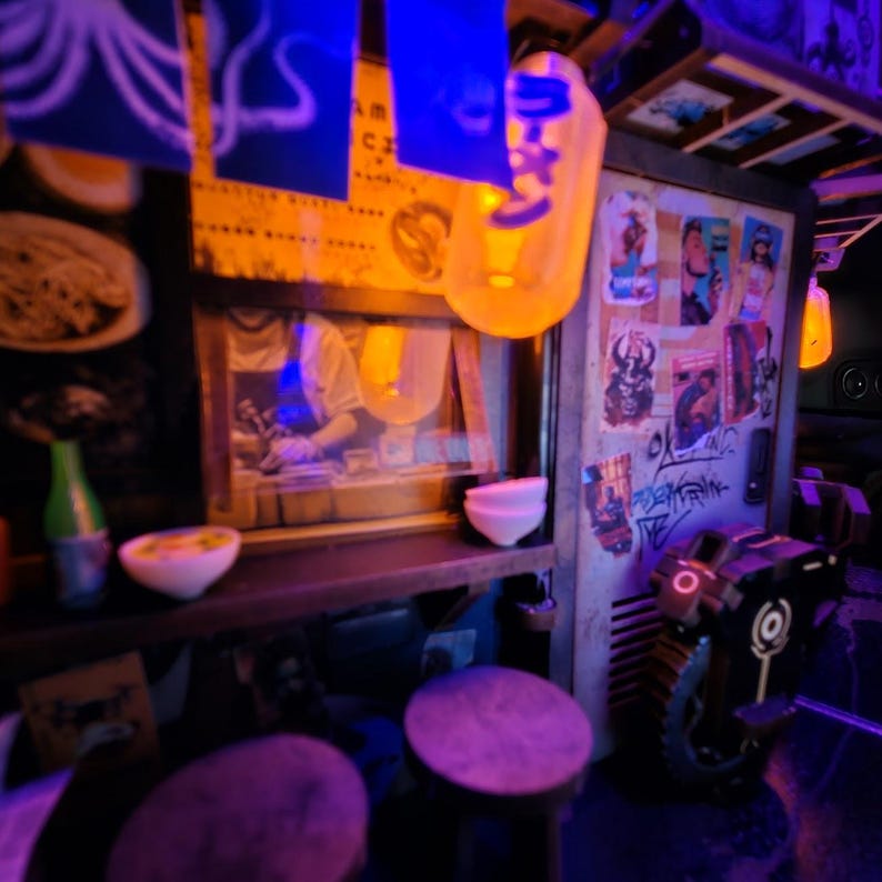 Op de afbeelding: Een miniatuur Japanse ramenwinkelsc&egrave;ne met een warme, uitnodigende gloed. De sc&egrave;ne omvat een kleine toonbank, krukken en posters. Er hangen lantaarns en een kleine robot. De winkel is versierd met Japanse karakters en voedselillustraties.