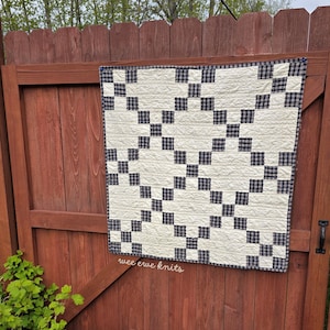 Könnte beinhalten: Ein quadratischer Quilt mit einem sich wiederholenden Muster aus cremeweißen Stoffblöcken und dunkel marineblau-weiß karierten Stoffquadraten. Der Quilt weist eine sichtbare Steppstruktur auf und der Text wee ewe knits ist darunter aufgedruckt.