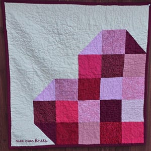 May include: A handmade quilt featuring a patchwork design in shades of pink and red, with a light green background. The quilt has a dark red border and the text "wee erwe knits" in the lower left corner.