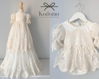 Puede incluir: Un vestido de bautizo de encaje blanco con falda larga y mangas abullonadas. El vestido está en un maniquí blanco y tiene una cinta de satén blanca alrededor de la cintura.