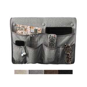 May include: A gray fabric bedside storage organizer featuring multiple pockets and three circular buttons along the top edge. The organizer holds a notebook, a clear water bottle, a smartphone with white earbuds, a remote control, and eyeglasses.