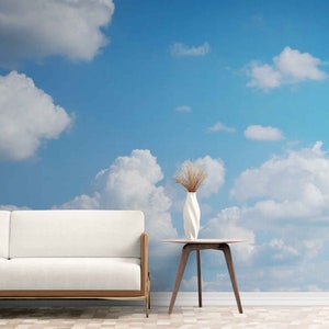 May include: A bright blue sky with fluffy white clouds serves as a backdrop for a modern living room. A white sofa with a wooden frame sits to the left, and a small round table with a white vase and dried plants is in the center.