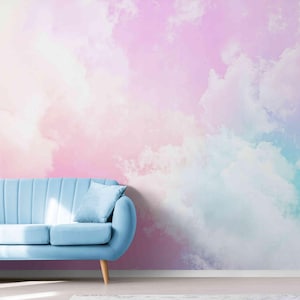 Op de afbeelding: Een lichtblauwe bank met twee kussens en een deken met franjes. De bank staat tegenover een muurschildering van een pastelkleurige lucht met roze, paarse en blauwe tinten. De kamer heeft een moderne, luchtige uitstraling.