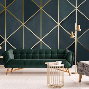 May include: A dark green velvet sofa with a gold metal frame sits in a living room with a dark blue and gold geometric patterned wallpaper. A gold metal side table with a white marble top sits in front of the sofa. A gray patterned armchair sits to the right of the sofa. Two gold floor lamps with white shades stand in the background.