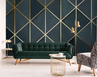 May include: A dark green velvet sofa with a gold metal frame sits in a living room with a dark blue and gold geometric patterned wallpaper. A gold metal side table with a white marble top sits in front of the sofa. A gray patterned armchair sits to the right of the sofa. Two gold floor lamps with white shades stand in the background.