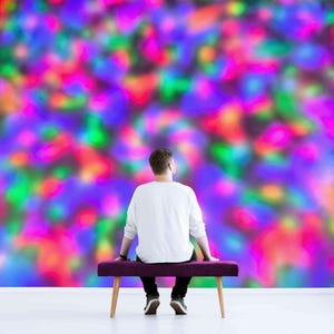 May include: A person sits on a purple bench in front of a wall with a colourful abstract design. The wall is covered in a blurred pattern of pink, blue, green, and yellow.