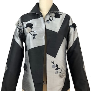 May include: A black and silver jacket with a geometric pattern and floral embellishments. The jacket has a collar and long sleeves.