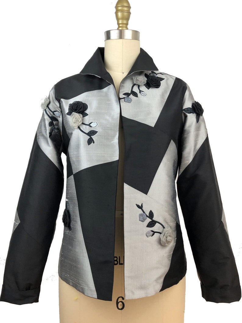 May include: A black and silver jacket with a geometric pattern and floral embellishments. The jacket has a collar and long sleeves.