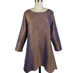 May include: A long-sleeved tunic featuring a shimmering iridescent finish that shifts between bronze and deep purple tones. The garment has a rounded neckline and a flared hemline that creates a subtle, flowing silhouette on the mannequin.