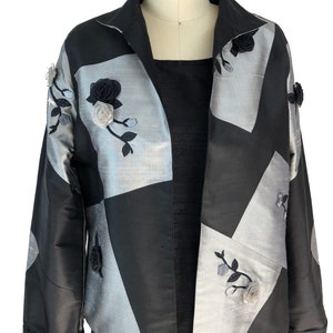 May include: A black and silver jacket with a geometric pattern and floral embroidery. The jacket has a collar and long sleeves.