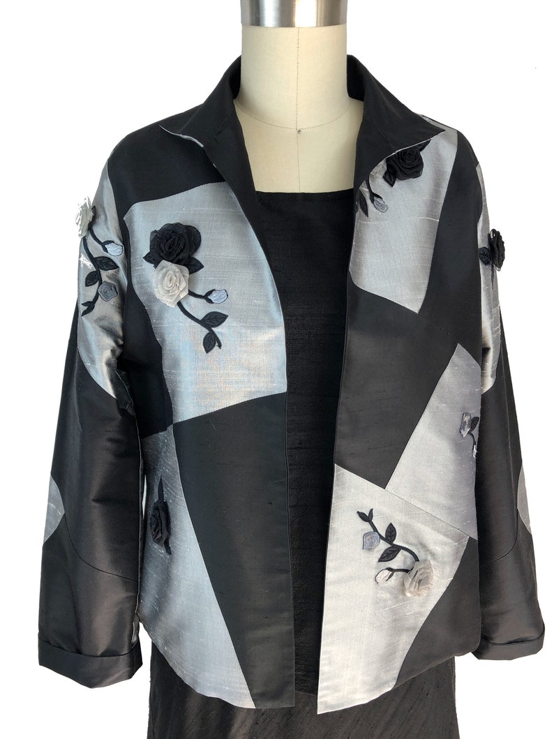 May include: A black and silver jacket with a geometric pattern and floral embroidery. The jacket has a collar and long sleeves.