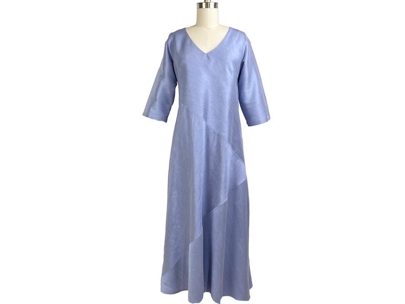 May include: A periwinkle blue midi dress featuring a V-neckline, three-quarter length sleeves, and a diagonal seam detail running across the front of the skirt. The garment is displayed on a mannequin and has a smooth, solid-colored fabric finish.