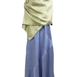 May include: A long, periwinkle blue dress with diagonal seam detailing, paired with a light sage green shawl draped over the shoulders. A beaded necklace with round, multi-colored stones rests at the neckline of the dress and shawl ensemble.