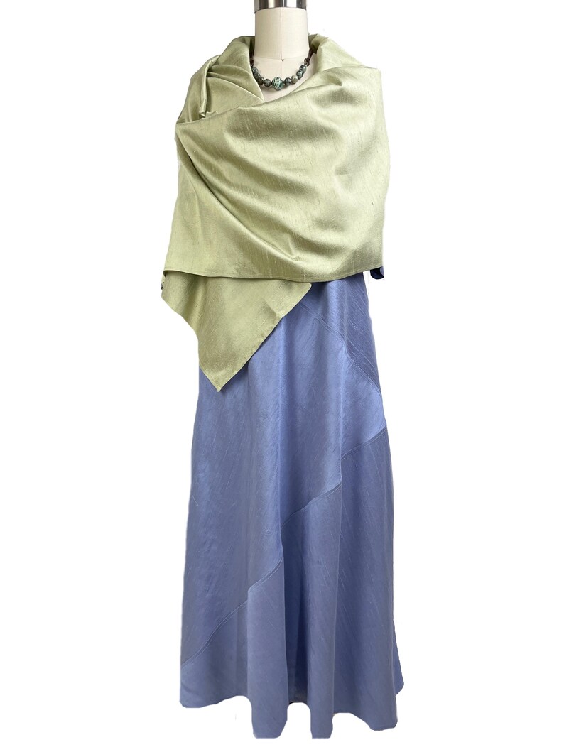May include: A long, periwinkle blue dress with diagonal seam detailing, paired with a light sage green shawl draped over the shoulders. A beaded necklace with round, multi-colored stones rests at the neckline of the dress and shawl ensemble.