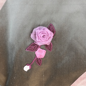K&ouml;nnte beinhalten: Eine rosa Stoffrose mit gr&uuml;nen Bl&auml;ttern ist auf einen dunkelbraunen Stoffhintergrund gen&auml;ht.