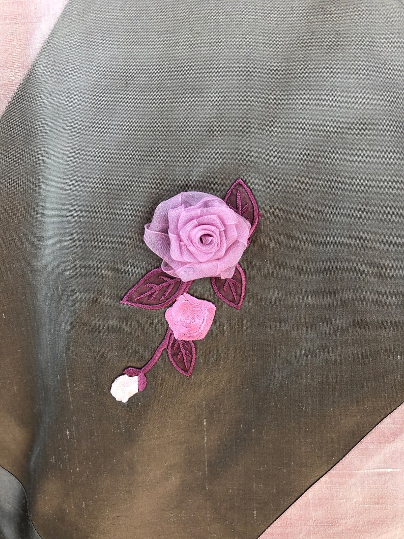 K&ouml;nnte beinhalten: Eine rosa Stoffrose mit gr&uuml;nen Bl&auml;ttern ist auf einen dunkelbraunen Stoffhintergrund gen&auml;ht.