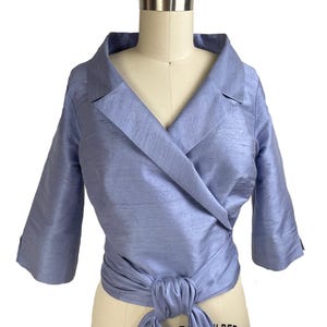 Può includere: Un top avvolgente azzurro con colletto stile blazer e maniche a tre quarti. Il top è legato in vita e ha un aspetto strutturato, probabilmente seta. L'indumento è esposto su un manichino.