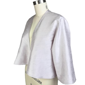 May include: A light purple, open-front jacket with wide sleeves. The jacket is made of a shiny, textured fabric and is displayed on a mannequin. The garment has a V-neckline and a cropped length.