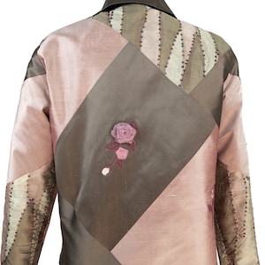 K&ouml;nnte beinhalten: Eine braune und rosafarbene gesteppte Jacke mit einer floralen Verzierung auf der R&uuml;ckseite. Die Jacke hat einen Kragen und lange &Auml;rmel.