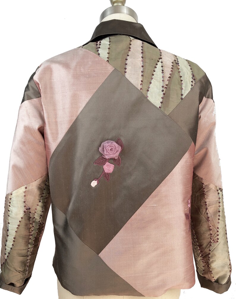 K&ouml;nnte beinhalten: Eine braune und rosafarbene gesteppte Jacke mit einer floralen Verzierung auf der R&uuml;ckseite. Die Jacke hat einen Kragen und lange &Auml;rmel.