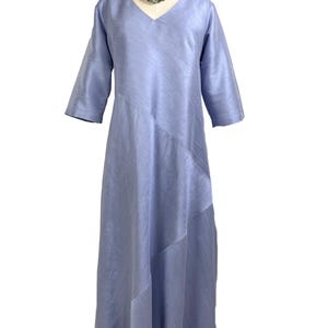 May include: A periwinkle blue midi dress with a V-neckline and three-quarter length sleeves. The garment features a subtle diagonal seam detail across the front, creating a layered effect in the fabric. The dress is displayed on a mannequin.