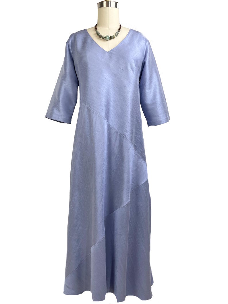 May include: A periwinkle blue midi dress with a V-neckline and three-quarter length sleeves. The garment features a subtle diagonal seam detail across the front, creating a layered effect in the fabric. The dress is displayed on a mannequin.