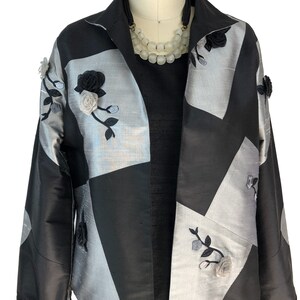 May include: A black and silver jacket with a geometric pattern and floral embroidery. The jacket has a collar and long sleeves.