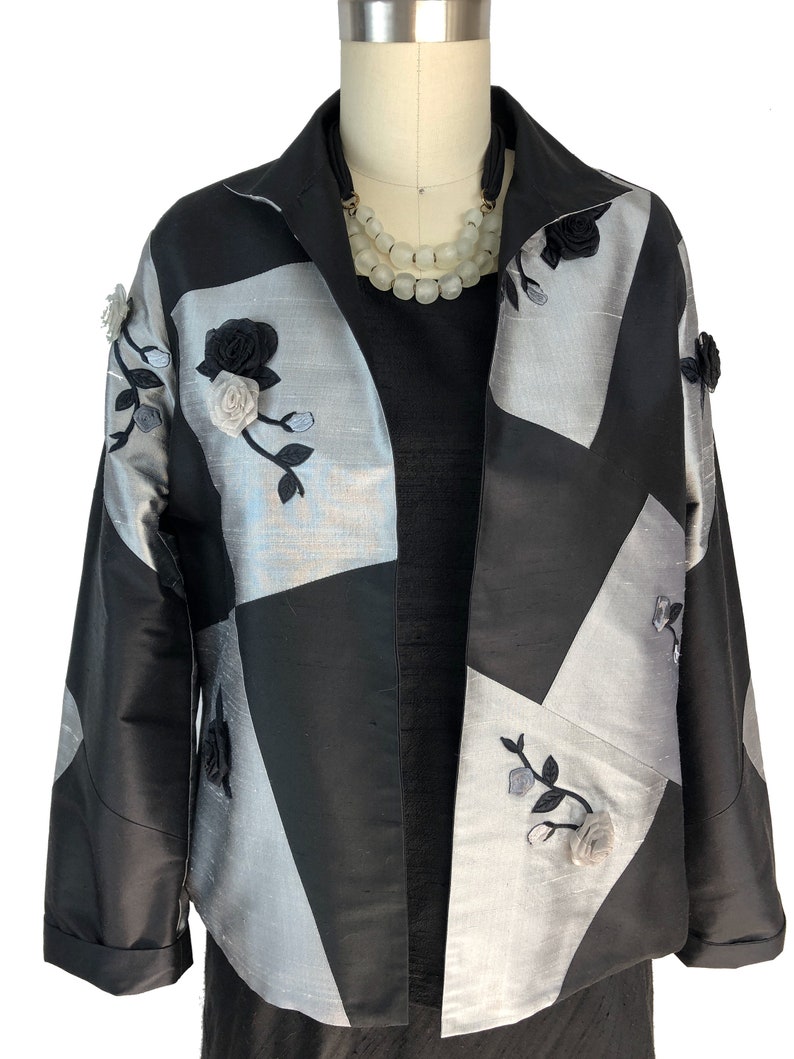 May include: A black and silver jacket with a geometric pattern and floral embroidery. The jacket has a collar and long sleeves.