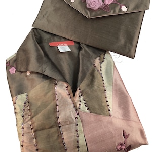 K&ouml;nnte beinhalten: Eine braune Seidenbluse mit einem rosa-gr&uuml;nen Blumendesign und eine passende braune Seidenclutch mit einem rosa Blumendesign. Die Bluse hat einen Kragen und einen Knopfverschluss. Die Clutch hat einen Klappenverschluss. Das Etikett "Lisbeth" ist auf der Bluse sichtbar.