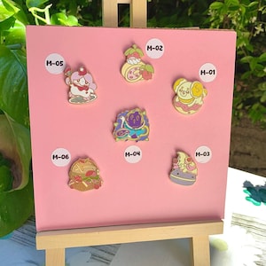 May include: A pink display board showcasing six enamel pins with vibrant dessert-themed designs. Each pin is labelled with a white sticker and a number from M-01 to M-06. The pins depict a dog, a cake, a fruit tart, and other sweet treats.